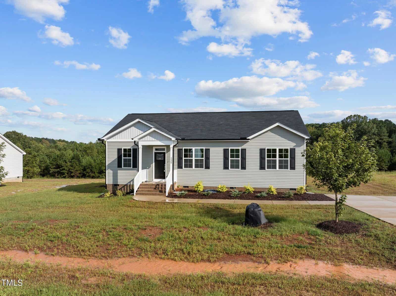 A single-story, light gray house with white trim and dark roof sits on a grassy lot with a small tree, driveway, and a landscaped front yard under a partly cloudy sky.