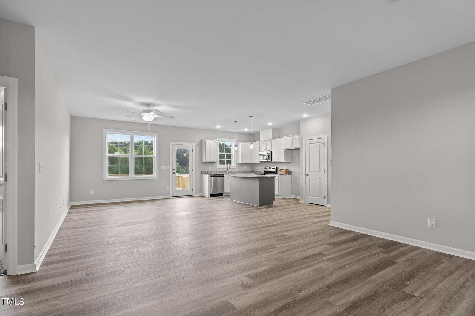 Open-concept room with wood-look flooring, white walls, a kitchen with island, white cabinets, stainless steel appliances, a ceiling fan, and large window. Glass door leads outside; space is unfurnished and well-lit.