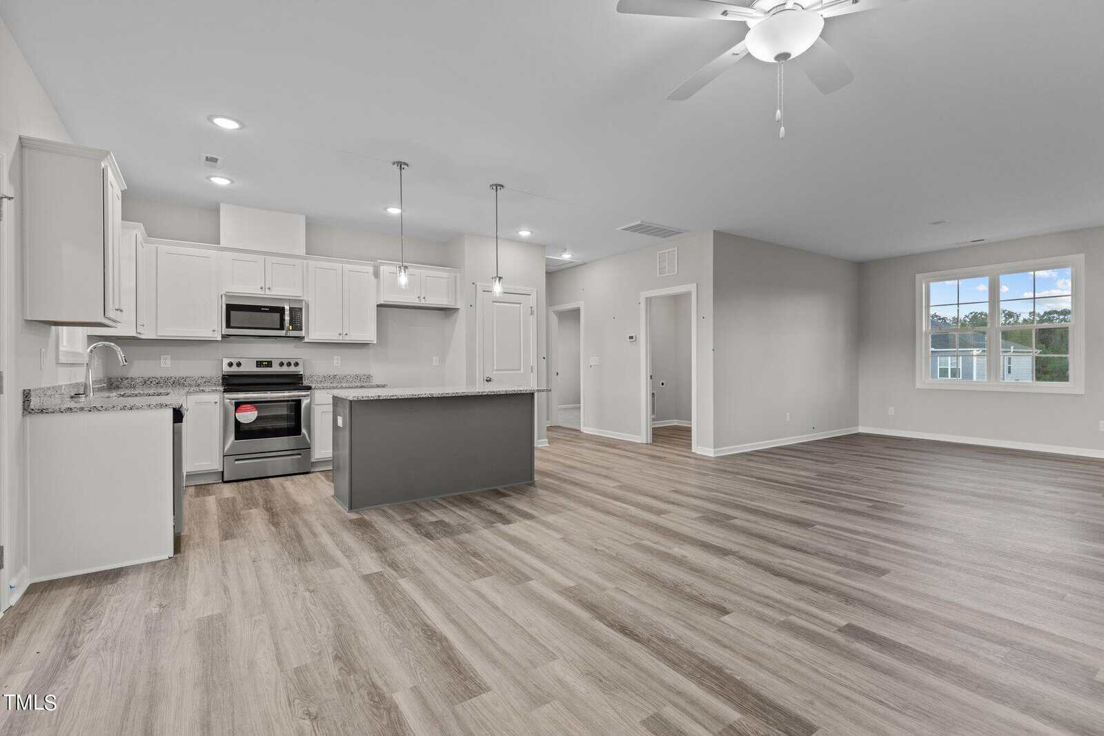 Spacious modern kitchen and living area with light gray cabinets, stainless steel appliances, granite countertops, pendant lighting, wood-look flooring, and large windows letting in natural light.