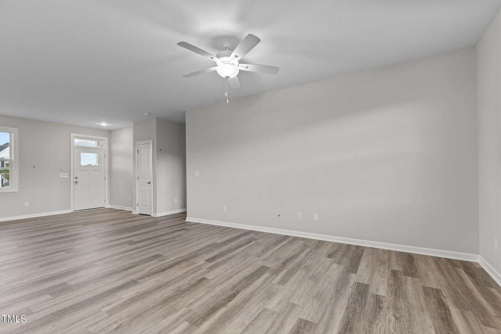 Empty room with light gray walls, wood-look flooring, a white ceiling fan with light, white trim, and two doors on the left wall. A small window is also visible near the entry door.