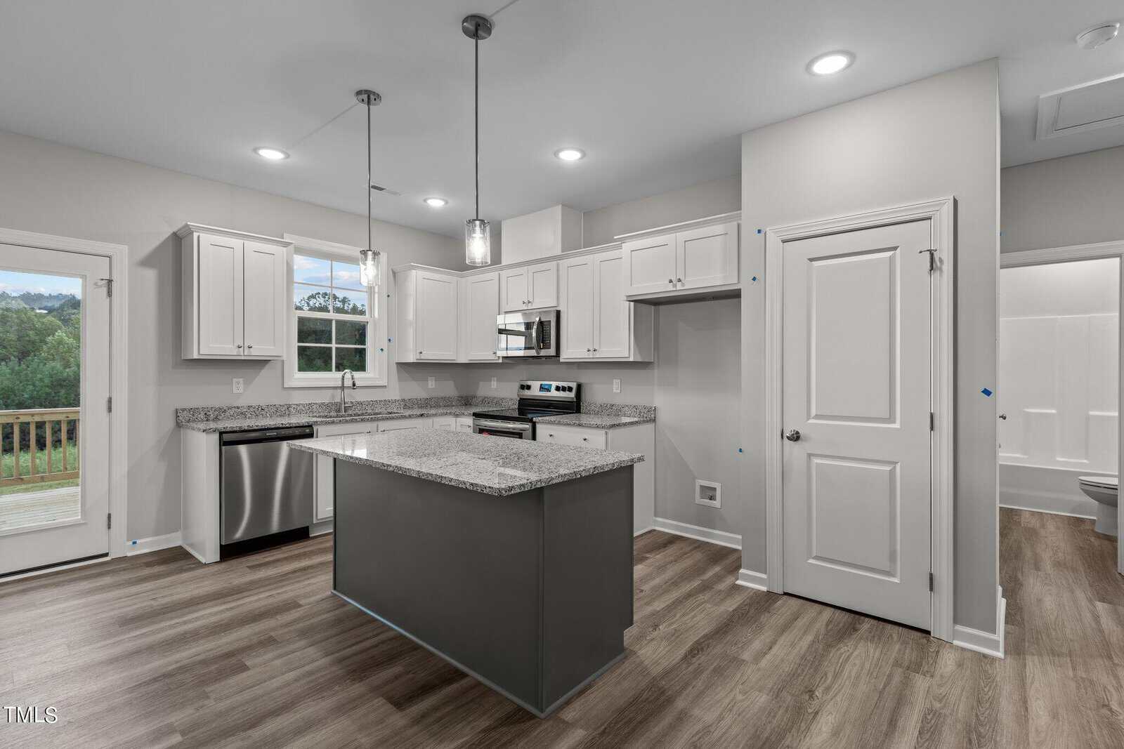 Modern kitchen with white cabinets, gray island, granite countertops, stainless steel appliances, pendant lights, wood flooring, and a door leading outside. A bathroom is visible through an open door on the right.