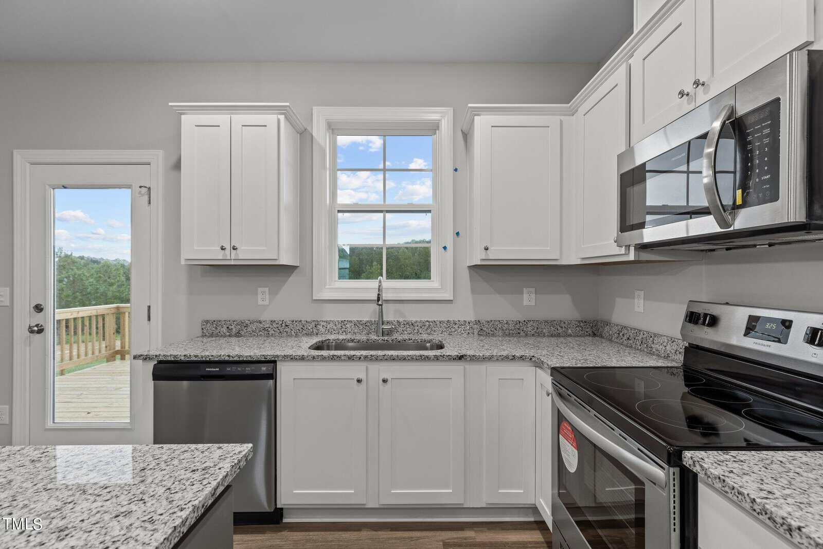 Modern kitchen with white cabinets, stainless steel appliances, granite countertops, a window over the sink, and a door leading outside to a wooden deck with a view of greenery and blue sky.