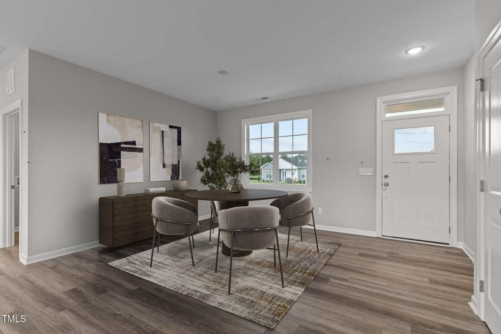 A modern dining area with four gray chairs around a round table on a rug, a sideboard with abstract art above it, and a door with windows letting in natural light.