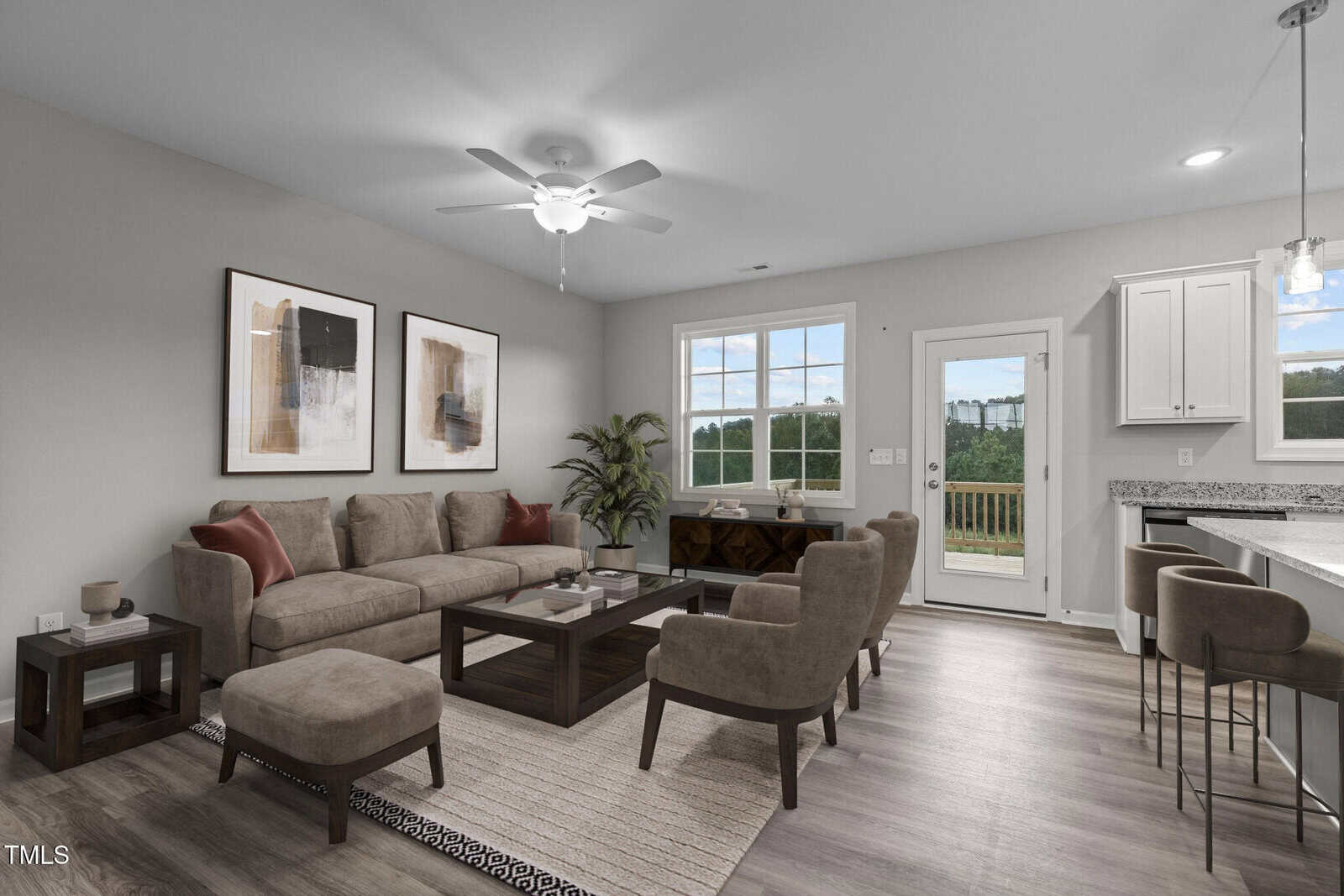 Modern living room with a neutral color palette, featuring a gray sectional sofa, two brown armchairs, a coffee table, wall art, a large window, and a door leading outside. Kitchen with bar stools is visible on the right.