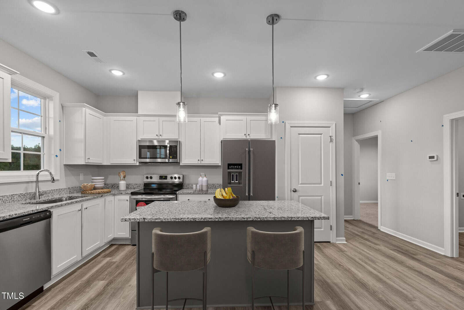 Modern kitchen with white cabinets, stainless steel appliances, granite countertops, and a large island with two barstools. Two pendant lights hang above the island. The flooring is light wood, and there is a window above the sink.