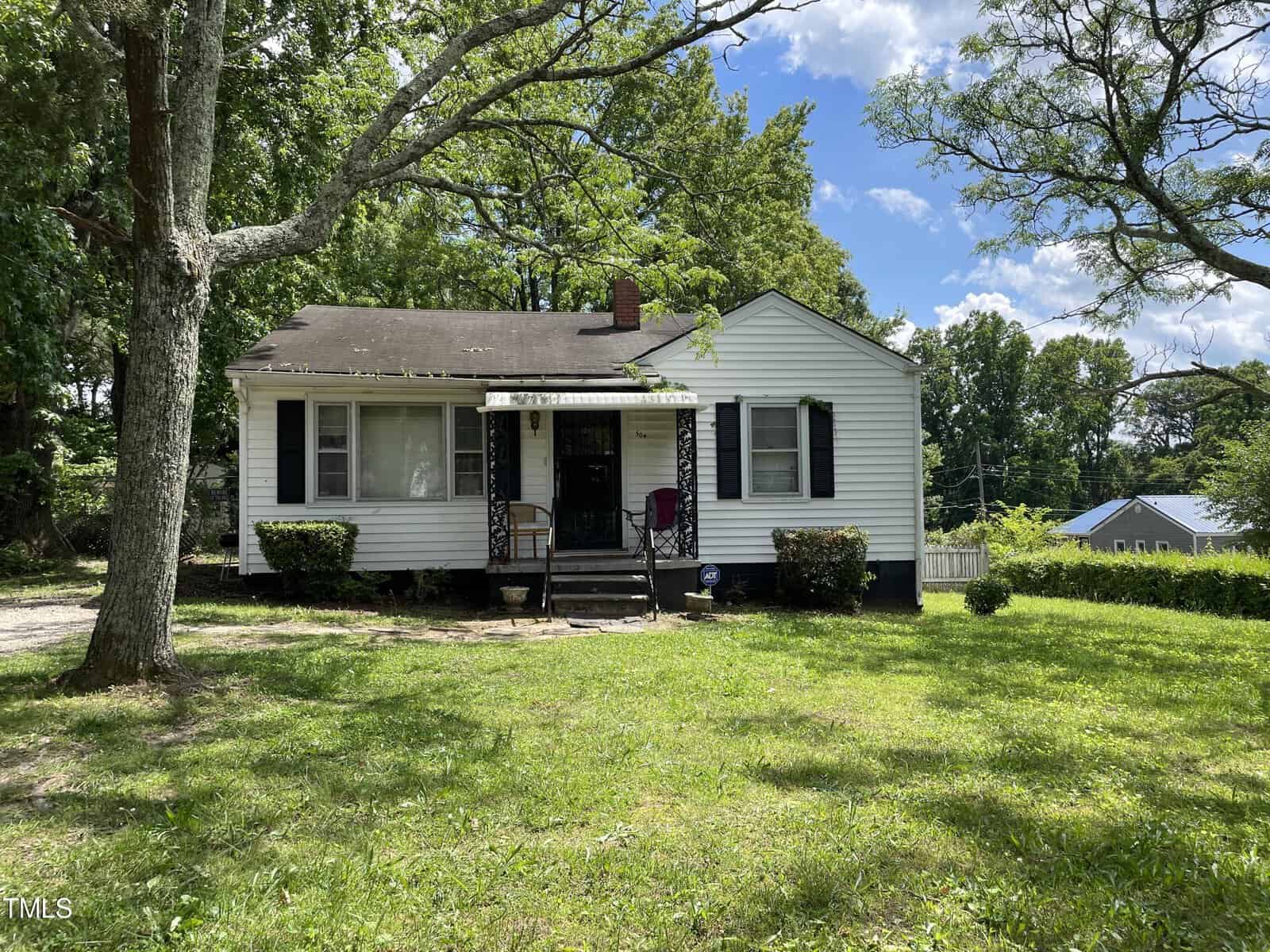 A small single-story white house with black shutters located in a lush, green yard at 504 Powell Street in Henderson, NC. The house features a covered front porch with a wooden railing, a few steps leading to the front door, and several trees providing shade. The sky is blue with some clouds.