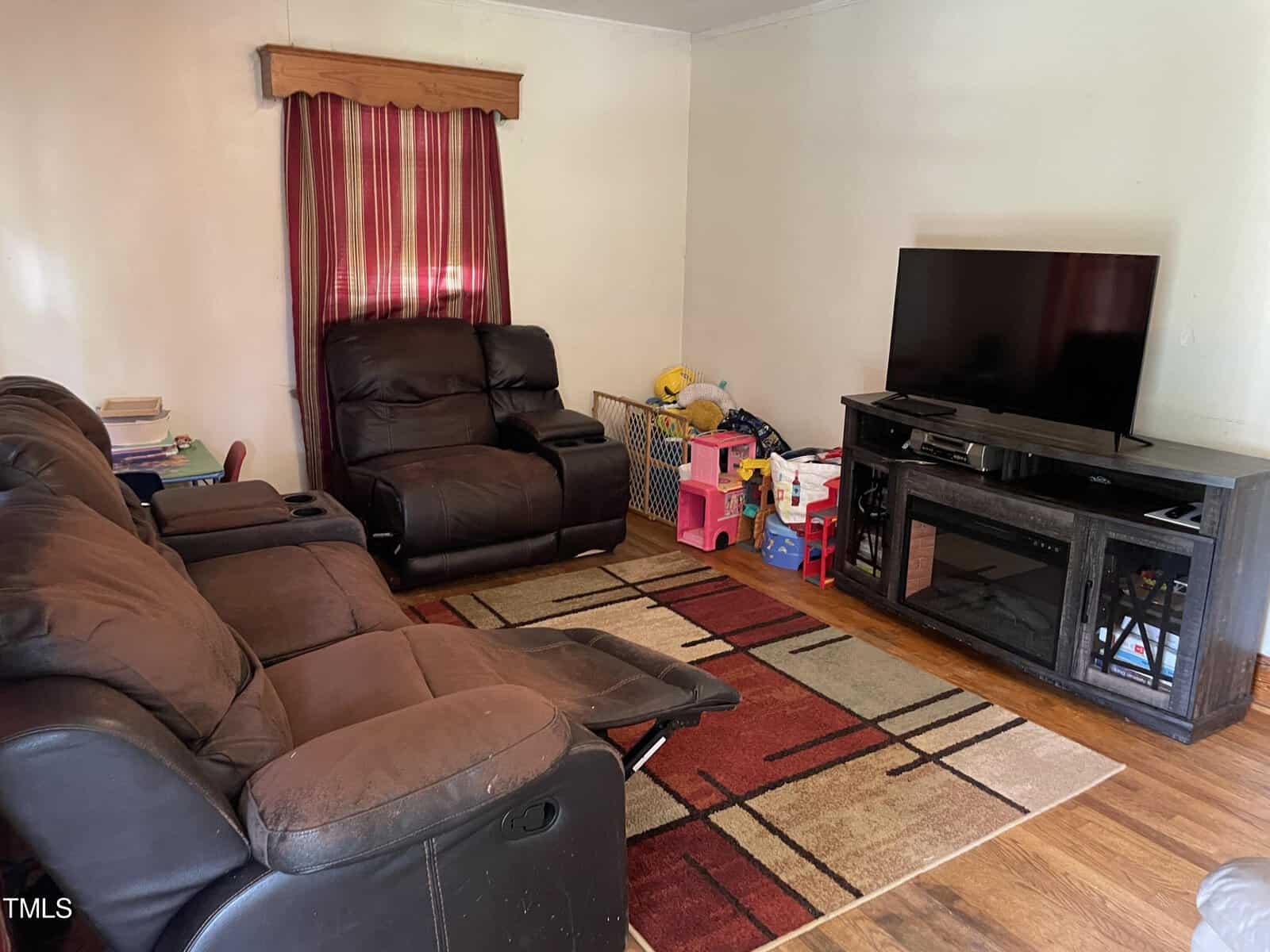 A living room with a large brown sectional sofa, a reclining armchair, and a flat-screen TV on a wooden stand. The room has a rectangular rug, striped curtain, and children's toys and books on the floor. Walls are plain, and the floor is hardwood.