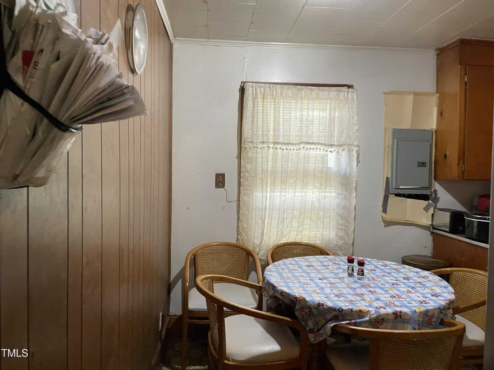 A small kitchen with wood-paneled walls, a round table with a patterned tablecloth, and four rattan chairs. A wall-mounted rack holds newspapers or mail. The window has sheer curtains, and the kitchen area includes an electrical panel and small appliances.