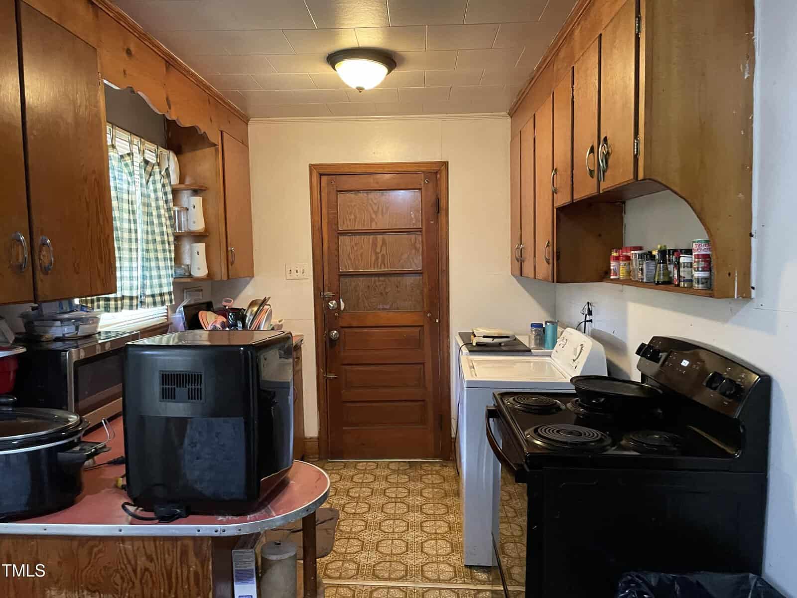 A small kitchen with wooden cabinets, a wooden door at the end, and white appliances, including a stove and a washing machine. A counter on the left holds various kitchen items, including a microwave and a slow cooker. The floor has a vintage patterned linoleum.