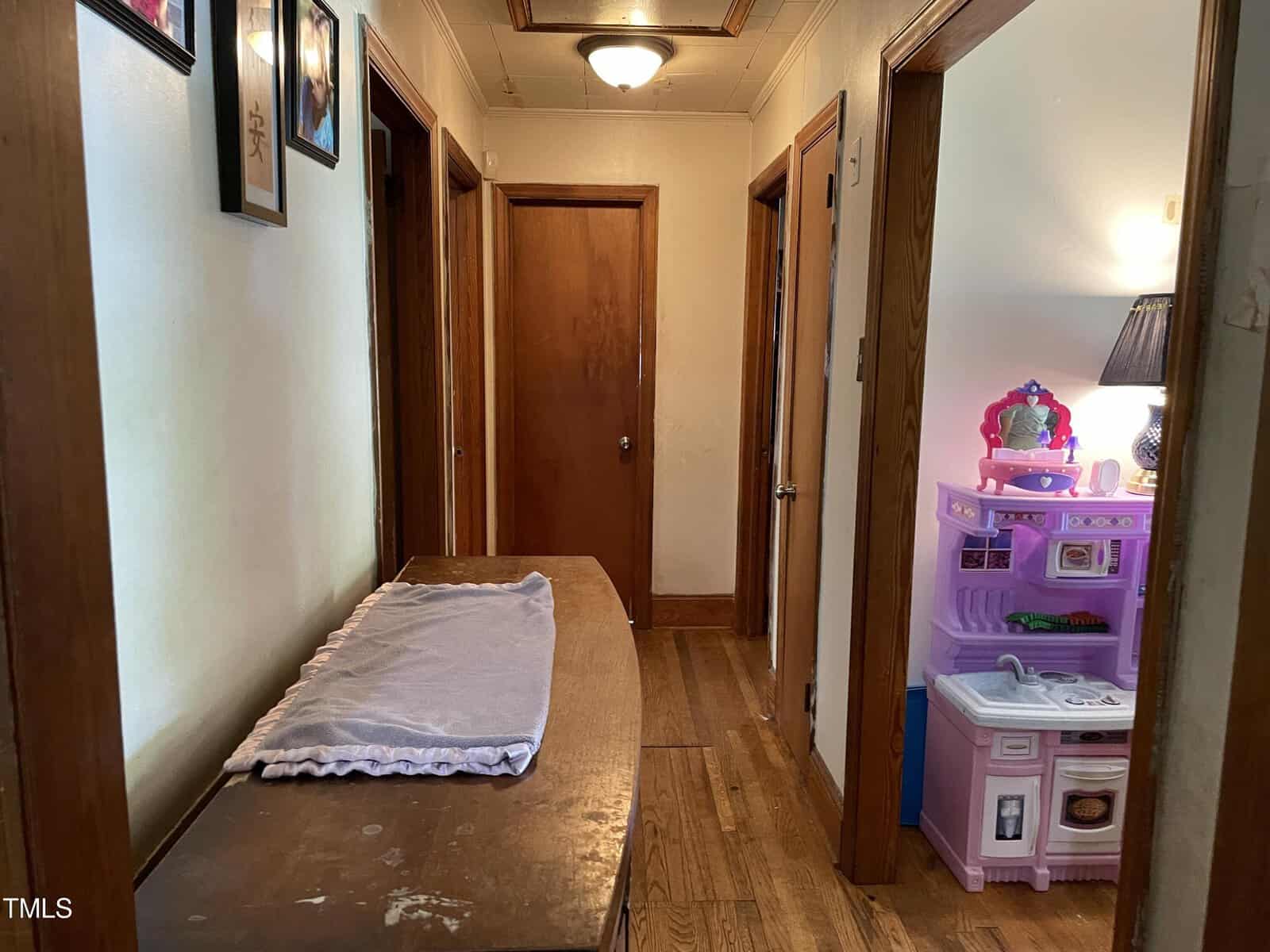 A hallway with wooden floors and doors. On the left, there's a dresser with a folded blanket. On the right, there is a child's pink and purple play kitchen set next to a lamp with a blue shade. The walls are decorated with framed pictures.