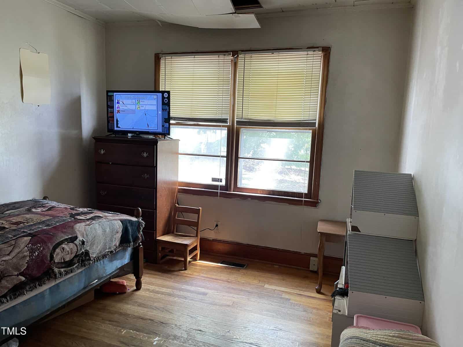 A modest room with a single bed, wooden dresser, and a small chair. The dresser has a flat-screen TV on top. The room has two windows with blinds, a simple wooden floor, and a dollhouse against the wall. The ceiling shows signs of damage.