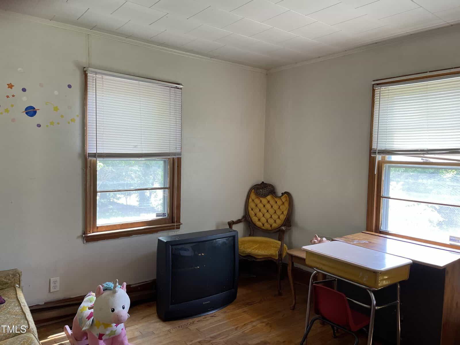 A small room with light-colored walls and hardwood floors. It features a vintage armchair, a CRT television on the floor, a children's table with two chairs, and a small plush unicorn toy in the corner. Two windows with blinds allow sunlight to enter.