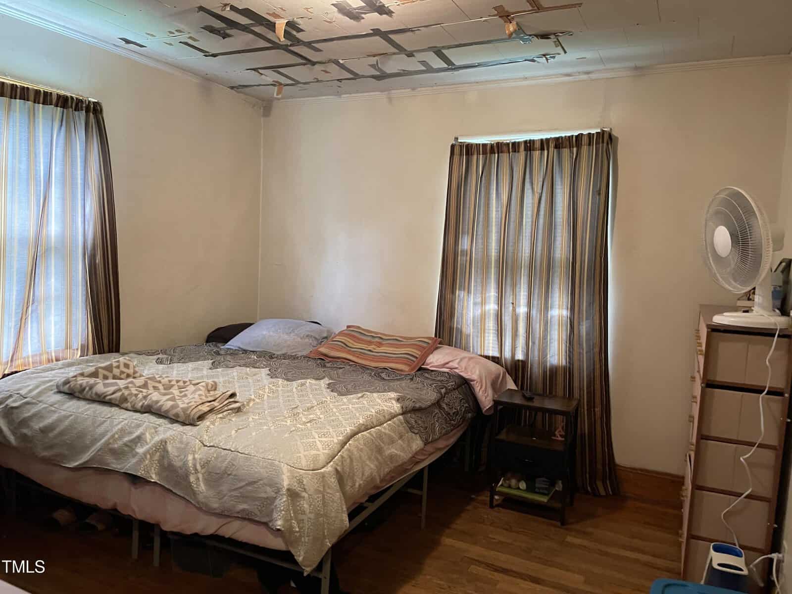 A bedroom with minimalist furnishings. A large bed with various blankets and pillows occupies most of the space. There are two windows with striped curtains, a fan on a dresser, and visible damage to the ceiling with tape patches. The walls are plain and light-colored.
