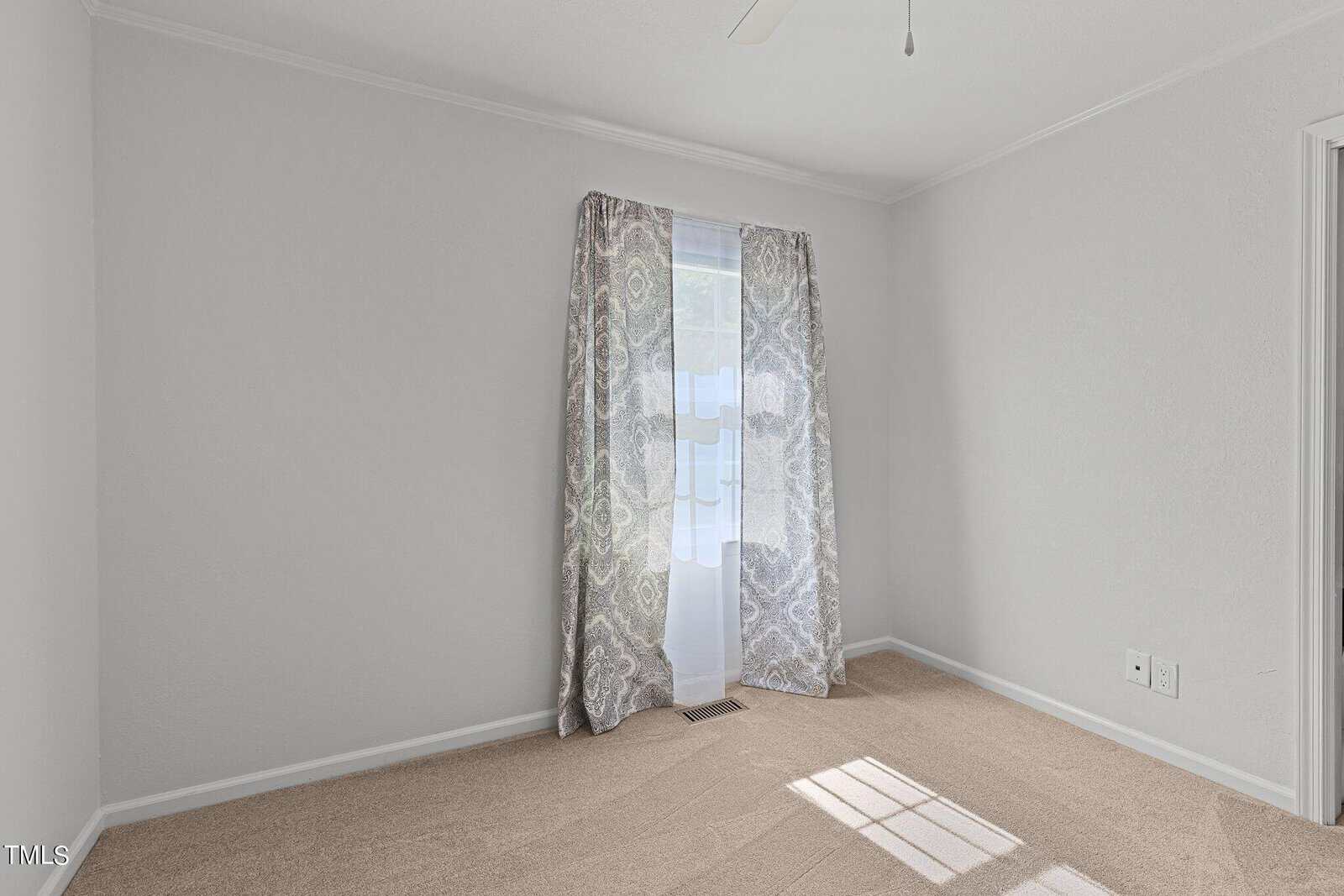 A small, empty room with white walls and beige carpet. Two patterned curtains frame a window that lets in natural light. A ceiling fan is partially visible at the top, and an electrical outlet is on the right wall.