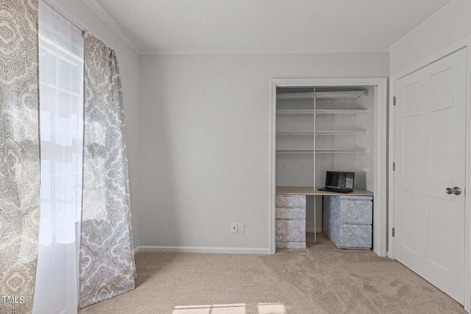 A small, empty room with light gray walls and beige carpet. The room features a window with patterned curtains and an open closet with white shelves and drawers. A black laptop is placed on one of the closet shelves.