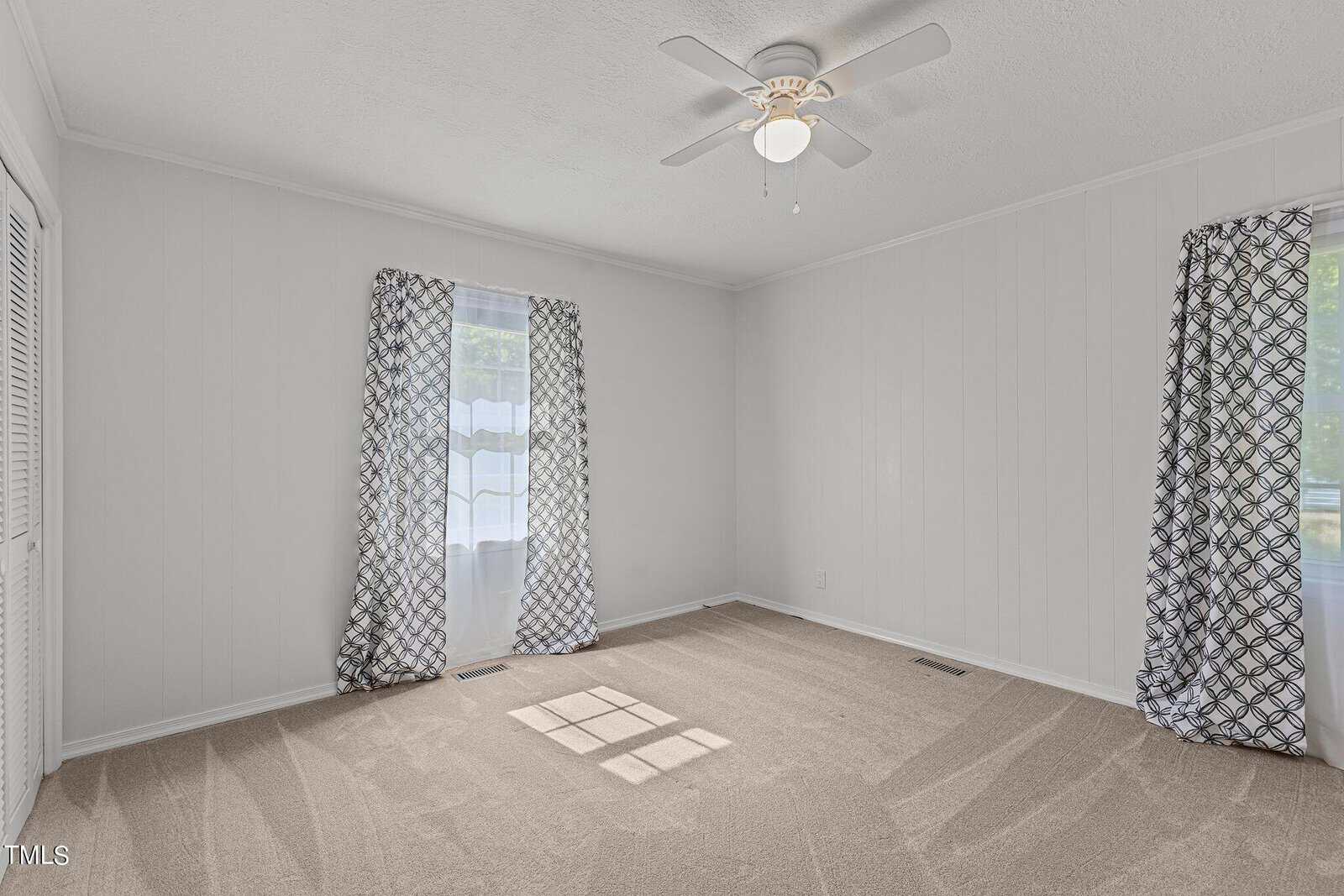 A small empty room with light-colored walls and beige carpet. There is a ceiling fan with a light in the center of the ceiling. Two windows with patterned curtains allow natural light to enter the room. The shadows of the window frames form squares on the carpet.