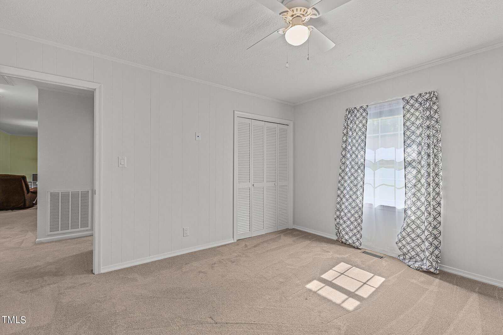 A small, empty room with beige carpet, white paneled walls, and a ceiling fan with a light fixture. The room has a closed white louvered closet door and a window with patterned curtains. An open door leads to another carpeted room.