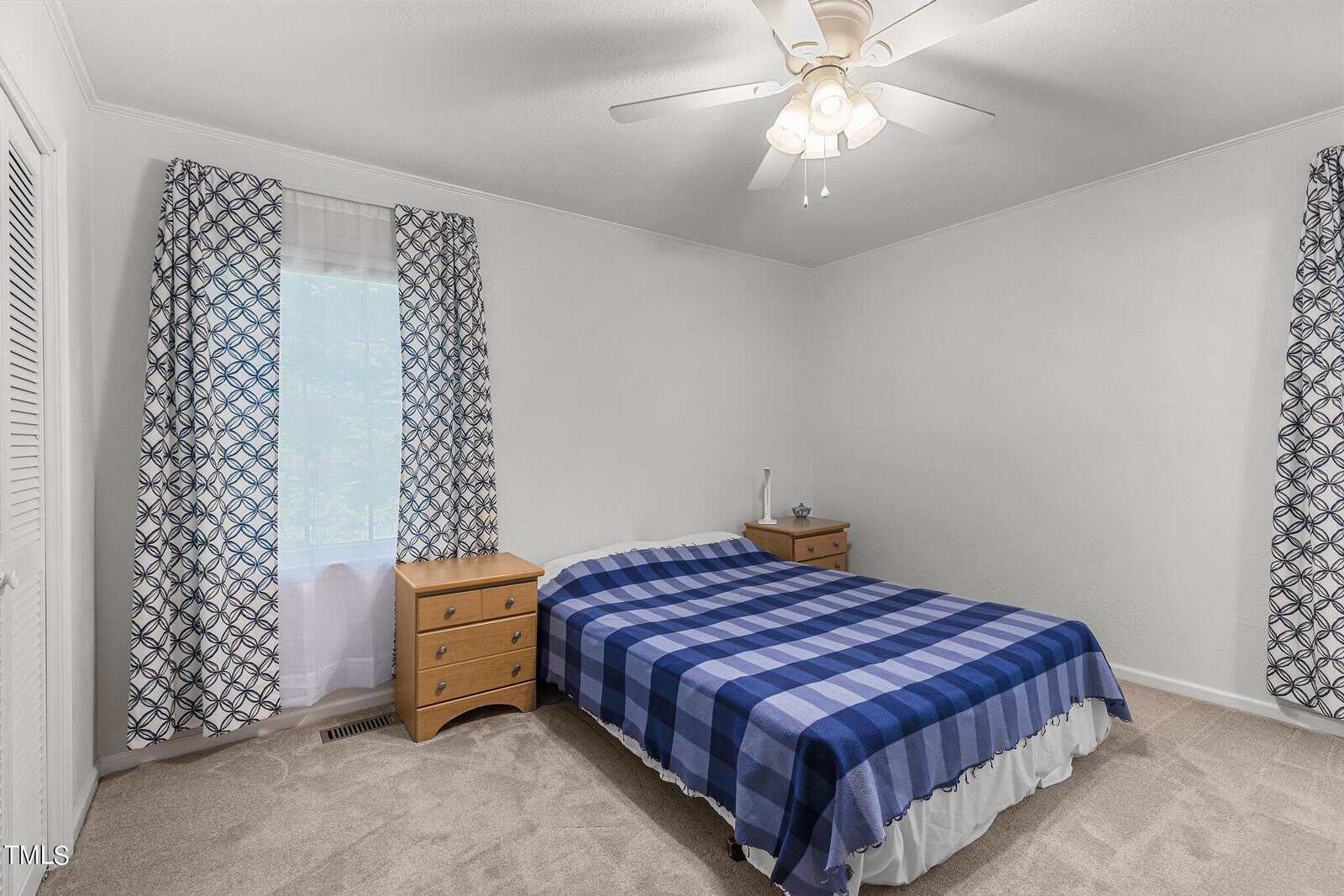 A small, well-lit bedroom with a double bed featuring a blue and white checkered blanket. The bed is flanked by two wooden bedside tables, each with a lamp. Patterned curtains adorn the windows, and a ceiling fan is positioned centrally above the bed.