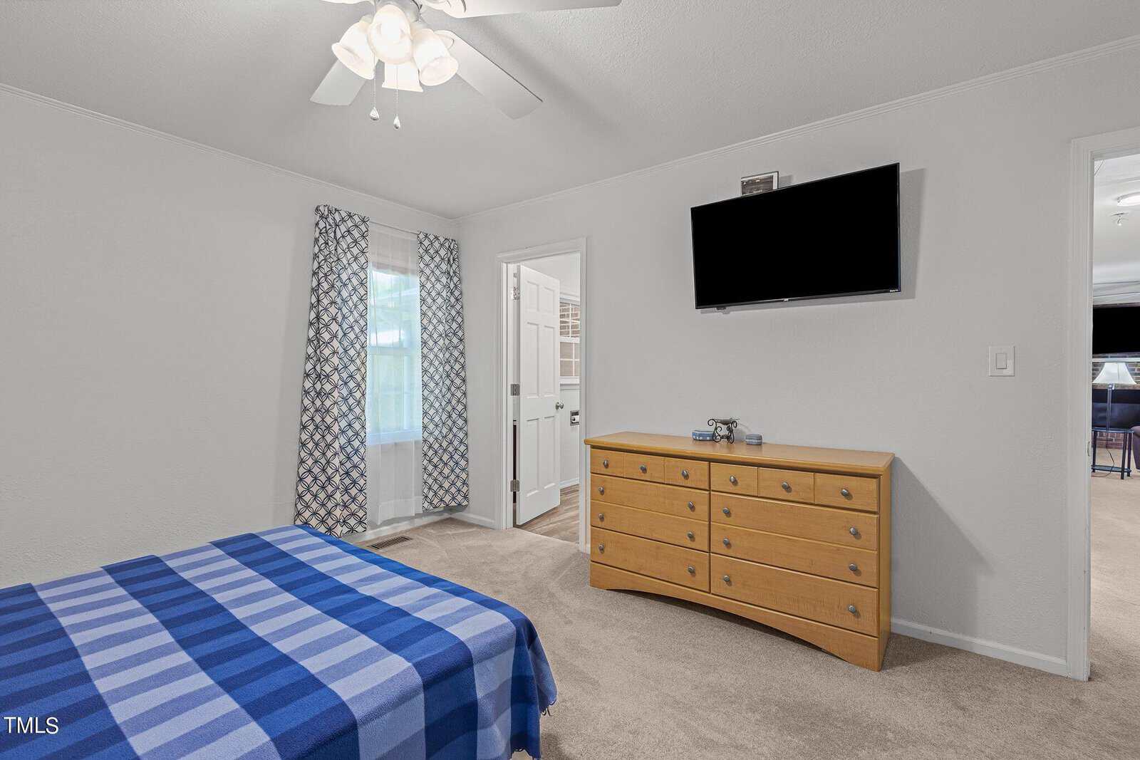 A bedroom with a blue and white checkered bedspread. Wooden dresser with a flat-screen TV mounted above it on the wall. Adjacent to the bed is a window with patterned curtains. An open door leads to an en-suite bathroom. Light ceiling fan above.