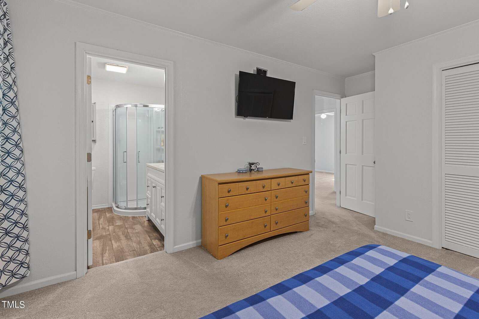 A bedroom with a blue checkered bedspread, a wooden dresser with a TV on the wall above it, and an open door leading to a bathroom with a shower enclosure. The room has carpeted floors and one window with blue-patterned curtains.