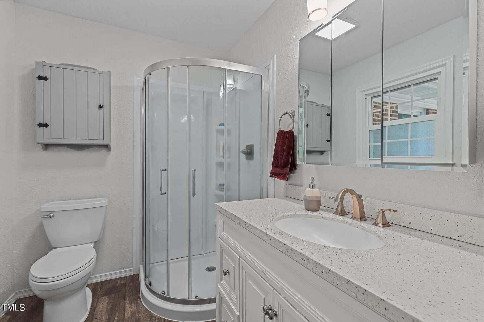 A modern bathroom featuring a curved glass shower enclosure, a white countertop vanity with a gold faucet and mirror above it. To the left is a toilet, and above it is a small gray wall cabinet. The floor is finished with dark wood.