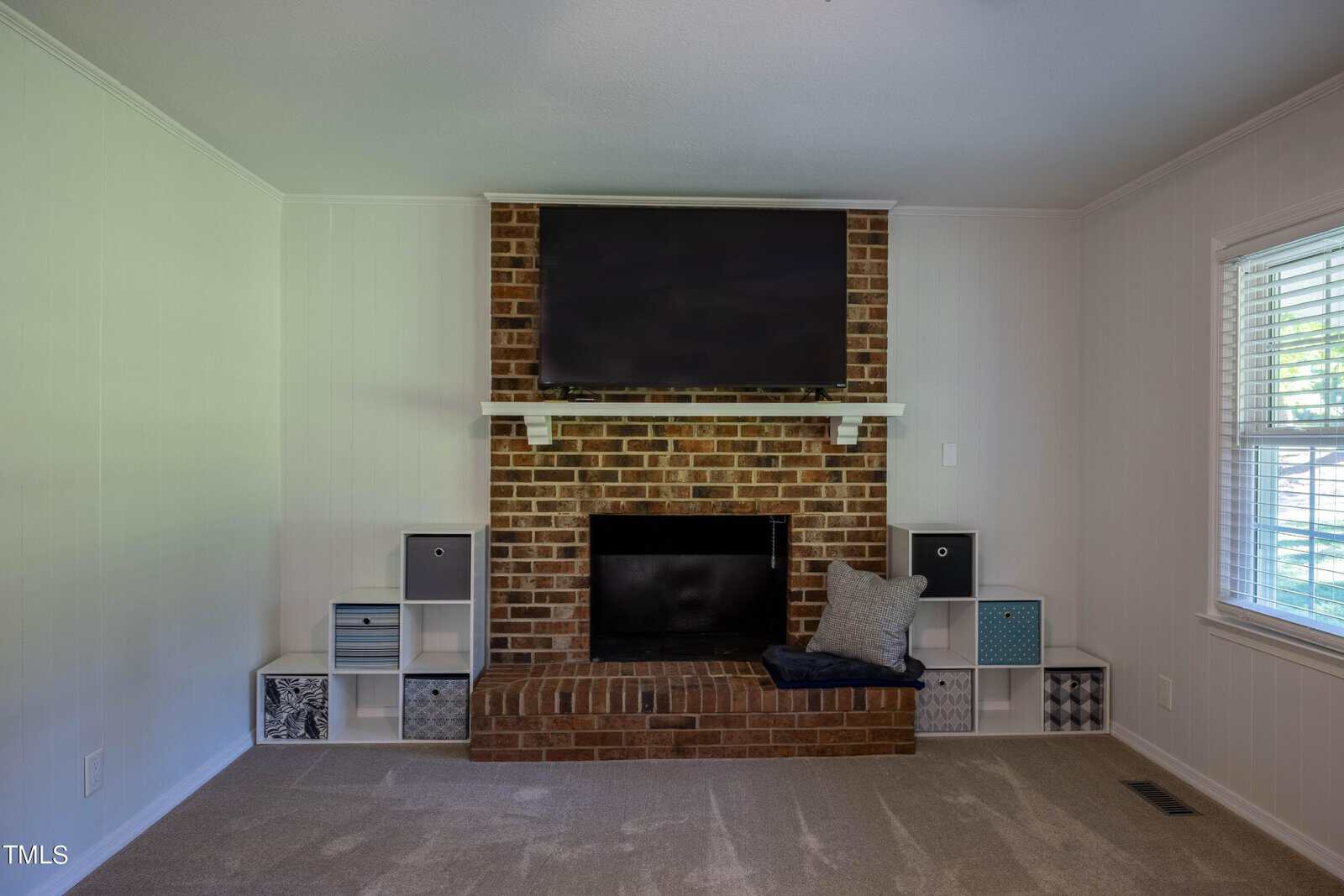A living room with a brick fireplace and a TV mounted above it. The fireplace hearth has a black screen and a gray cushion. Surrounding the fireplace are built-in white shelves with various storage bins. A large window with white blinds is on the right.