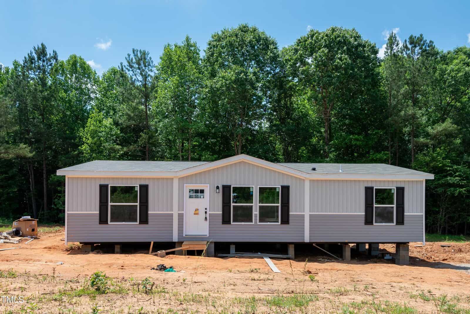 A single-story, pre-fabricated home at 533 Madison Grove Lane in Henderson, NC, sits on a dirt lot surrounded by lush green trees. The house features a gray and white exterior with multiple windows and a white door slightly left of center. The elevated foundation exposes wooden supports beneath the structure.