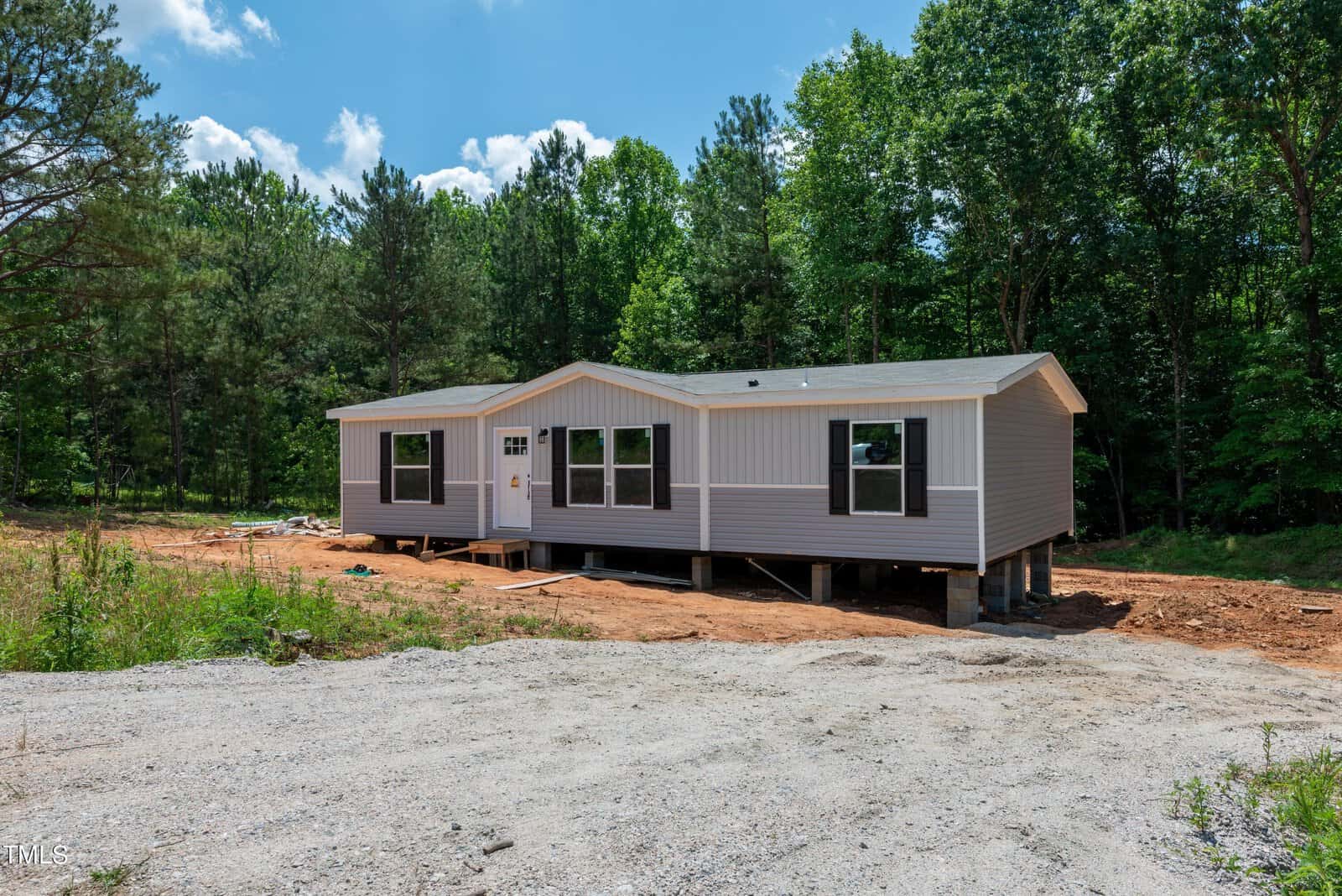 A newly constructed, single-story modular home with light gray siding and white trim sits elevated on stilts with visible wooden supports. The house is surrounded by dirt and greenery, with a forested area and blue sky in the background.
