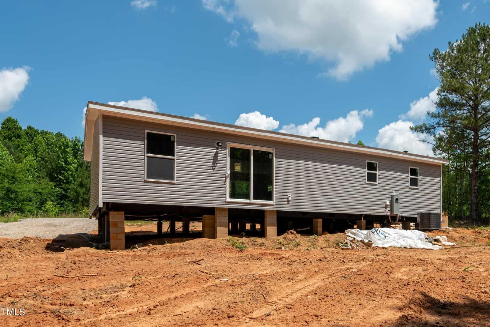 A single-story modular home with beige siding is elevated on wooden supports in a clearing surrounded by trees. The area features red dirt ground and some construction materials are visible on the right side. The sky is blue with fluffy clouds.