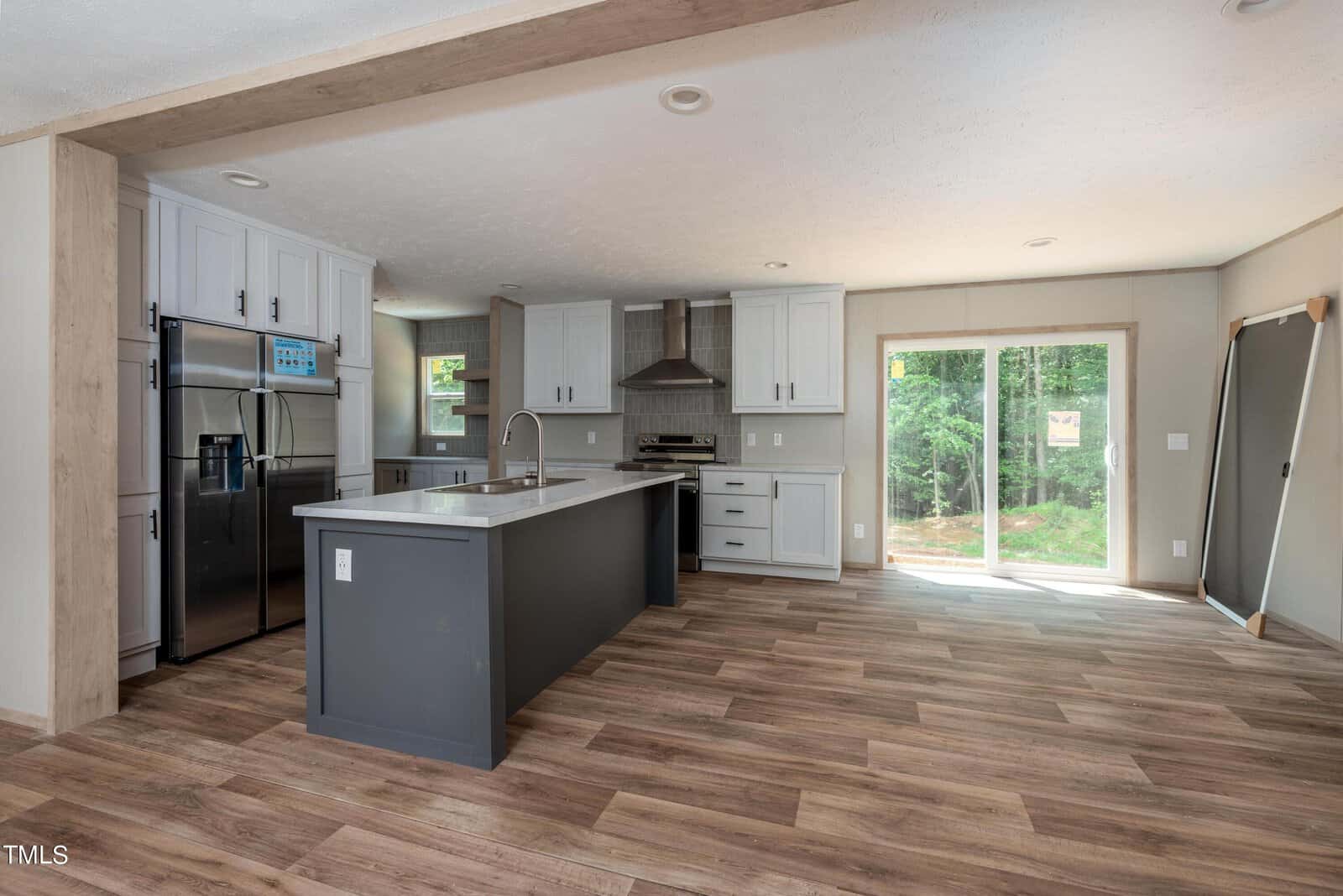 A modern kitchen with wooden flooring, white cabinets, a gray island with a sink, and stainless-steel appliances including a refrigerator and oven. Large sliding glass doors on the right allow natural light to enter, offering a view of the outdoors.