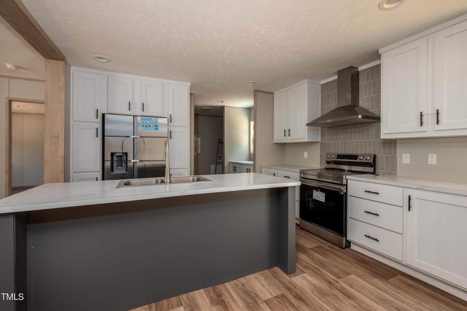 A modern kitchen with white cabinets, stainless steel appliances, and a large island with a gray base and white countertop. The kitchen features a dual sink, electric stove with a hood, and wood-patterned flooring. Sunlight filters in through a window.