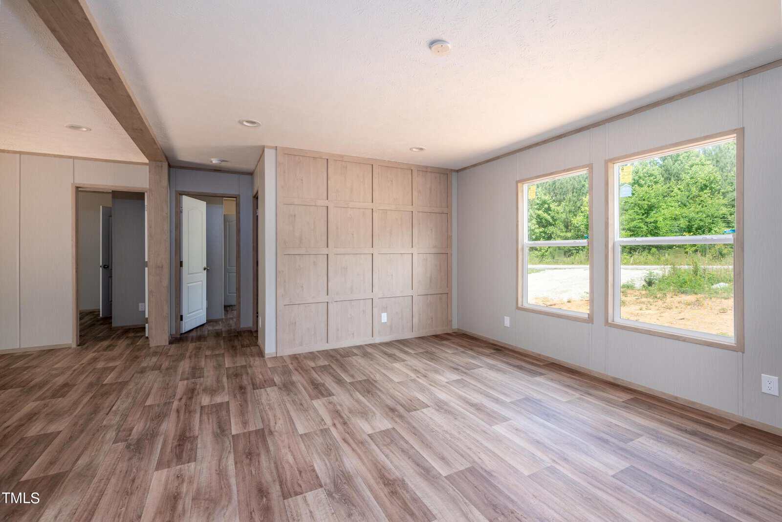 A spacious, unfurnished room with light wood-patterned flooring and neutral-toned walls. Two large windows on the right side let in natural light, showcasing a green outdoor view. An open doorway and an interior hallway are visible on the left.
