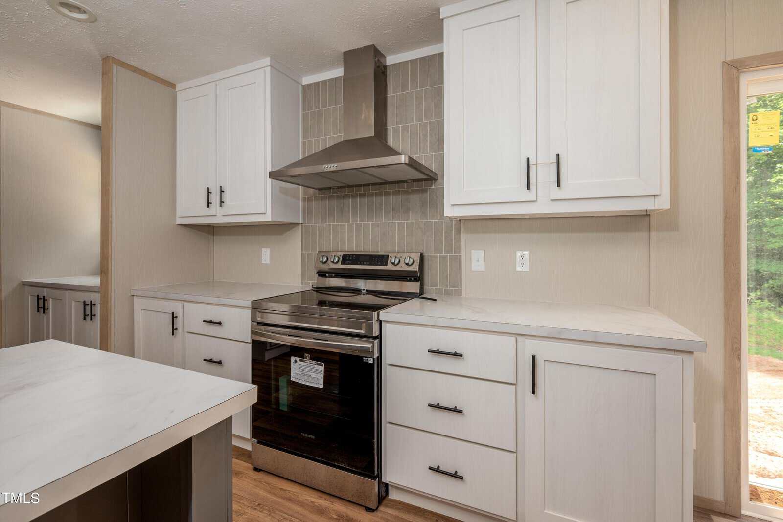 A modern kitchen with white cabinets, stainless steel appliances, and a large range hood above a stove. The backsplash features gray tiles, and the countertops are white. The floor is light-colored wood, and there is a window to the right allowing natural light in.