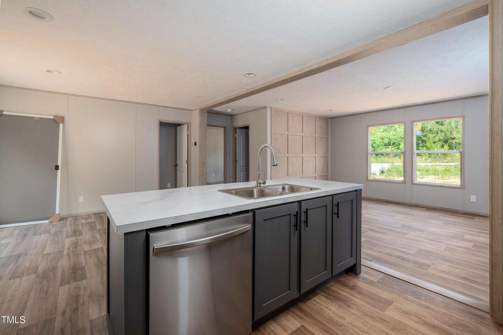 Modern open-concept kitchen and living area featuring a central kitchen island with a built-in sink and dishwasher, light wood floors, neutral wall tones, and large windows allowing natural light to fill the space. A door and hallway are visible in the background.
