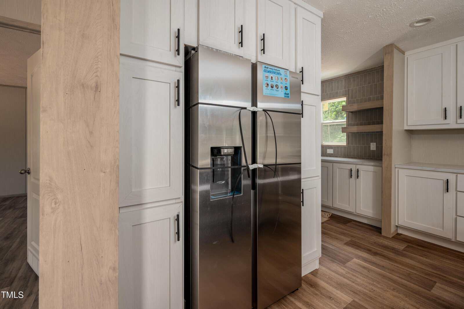 A modern kitchen features white cabinets, wood flooring, and a stainless steel refrigerator with a water dispenser and instruction sheet attached to the freezer door. A window in the background brings natural light into the space.