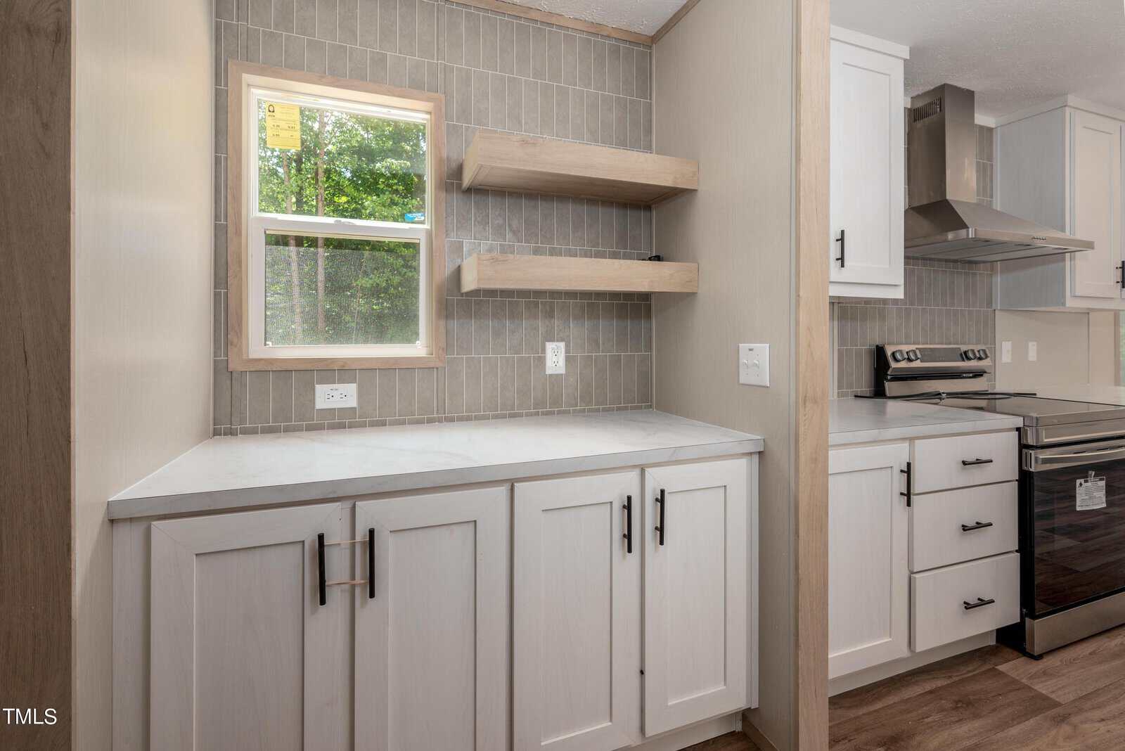 A modern kitchen with light gray tiled backsplash, white cabinets with black handles, and light wood open shelves. A window above the counter illuminates the space. The kitchen is equipped with a stainless steel stove and range hood. The countertop is white marble.
