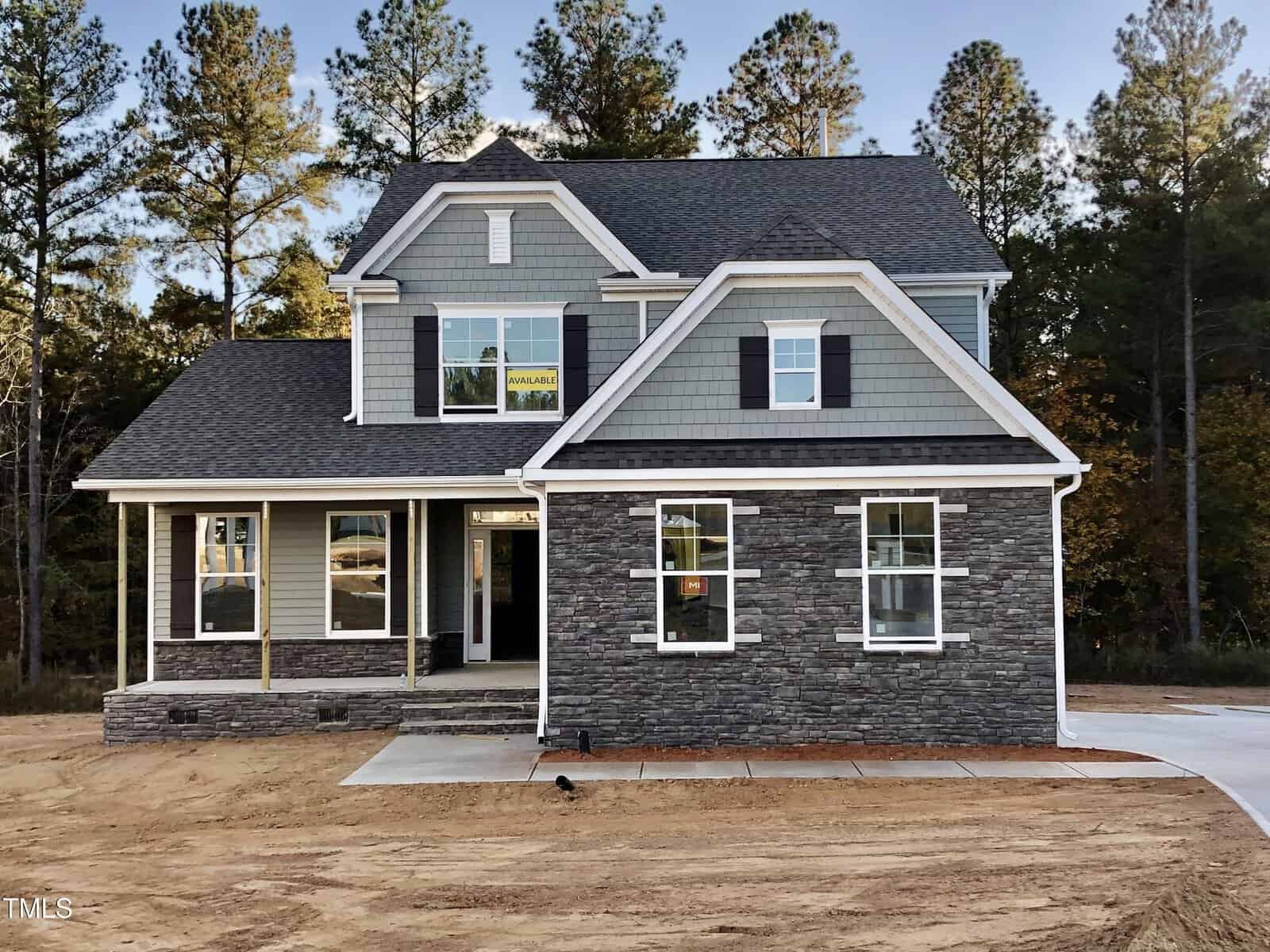 Two-story suburban house with gray siding and stone facade, white trim, black shutters, and a covered front porch. The yard is unlandscaped with bare earth, and tall pine trees are visible in the background.
