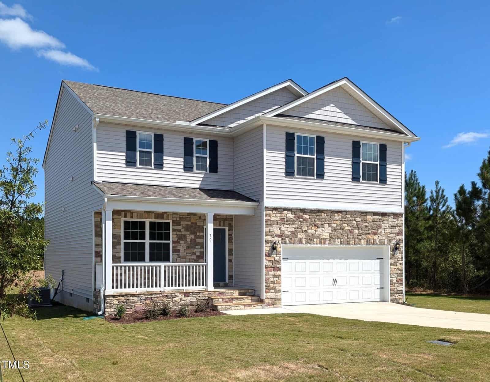 A two-story suburban house with light-colored siding, stone accents on the front, a covered porch, and a white double garage. The house sits on a lawn with a driveway and trees in the background under a blue sky.