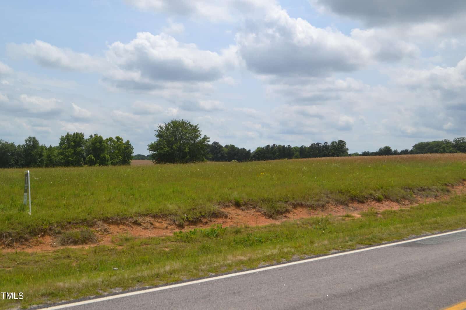 A rural landscape featuring an empty green field with a single tree in the center near the horizon. The sky is partly cloudy, and a paved road runs across the foreground. Additional trees are visible in the distance.