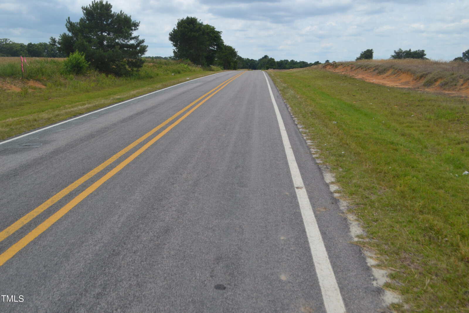 A rural road stretches into the distance under a partly cloudy sky. The road is lined with grass fields on both sides, with a few trees visible in the background. A double yellow line runs down the center of the road.