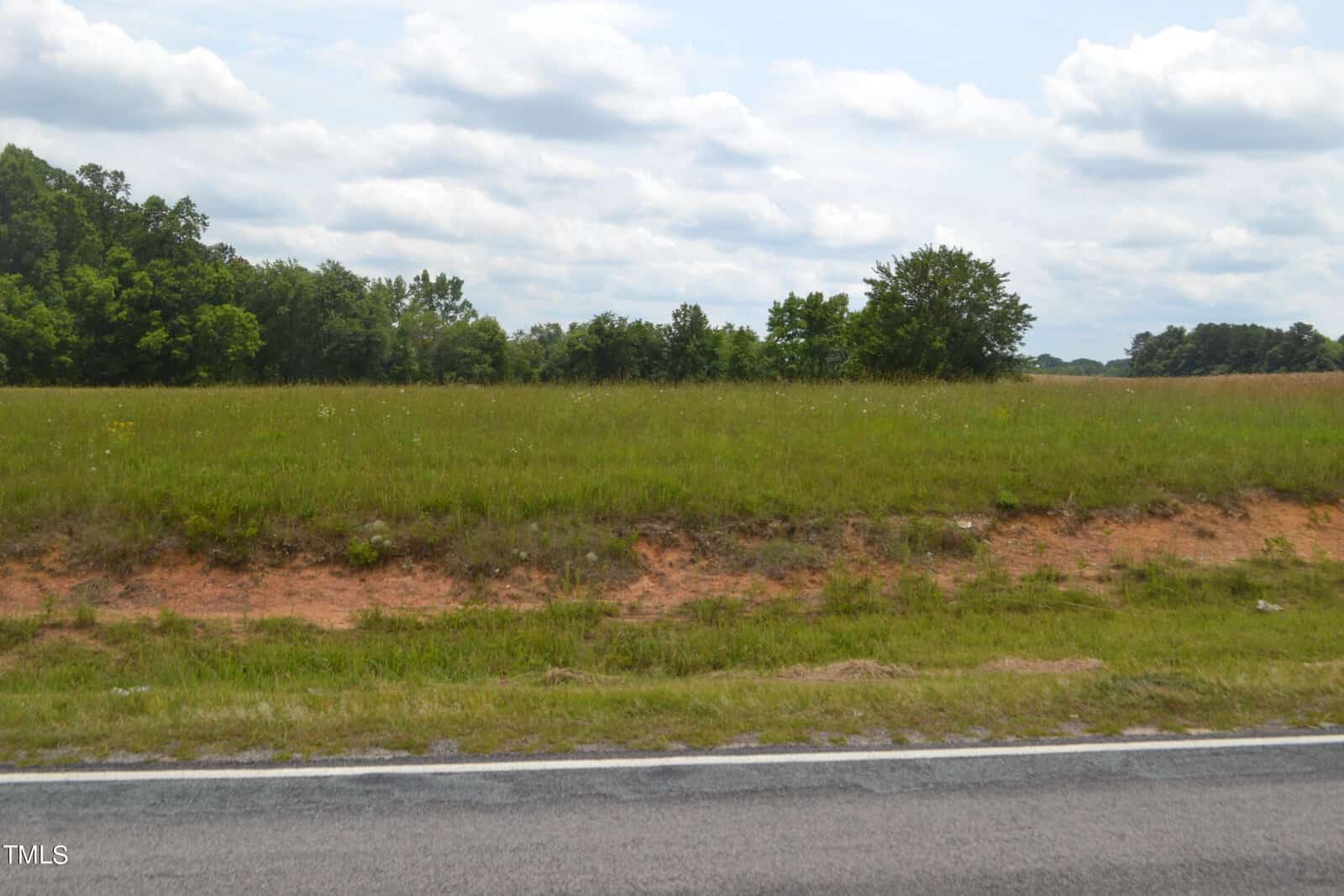 A gently sloping field with patches of green grass and reddish-brown soil is adjacent to a paved road. In the background, there are clusters of green trees under a partly cloudy sky.