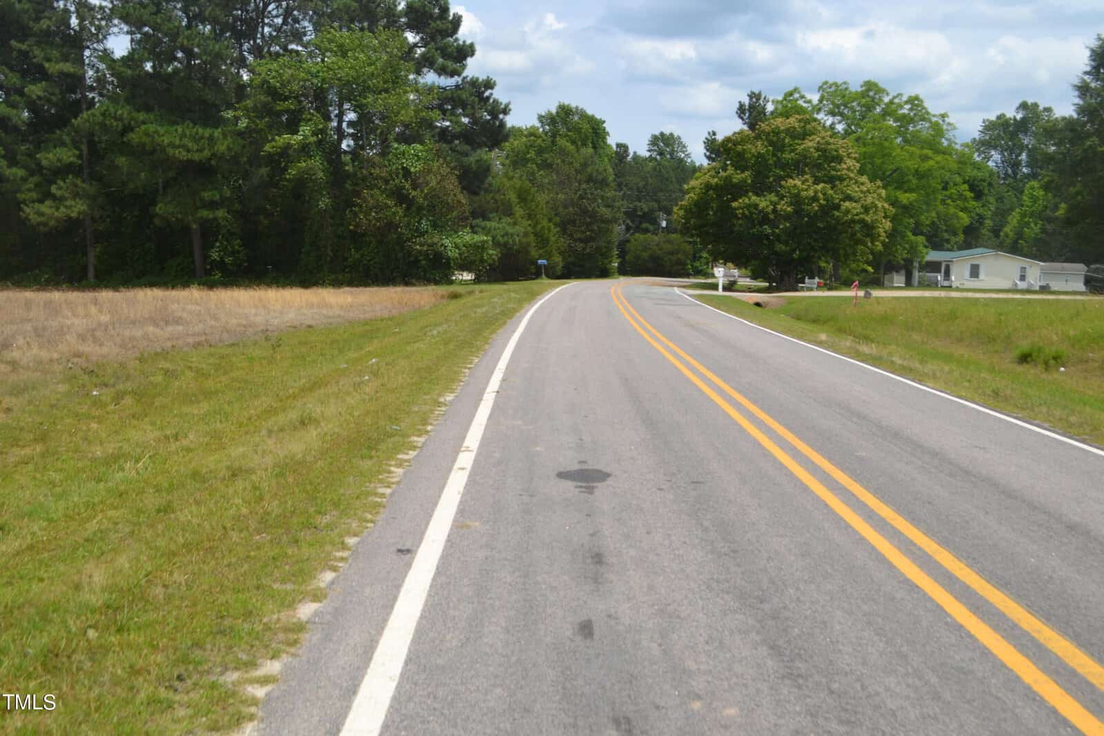 A paved, two-lane road with double yellow lines curves gently to the right. Trees and grass line the road on both sides, and a single house with a slanted roof is visible on the right among the green foliage under a partly cloudy sky.