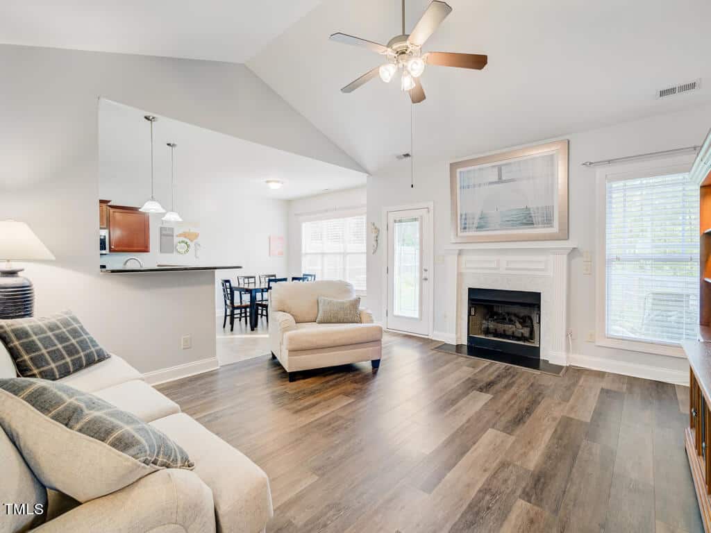 Spacious living room with wooden floors, beige sofa, armchair, and a fireplace with art above. Ceiling fan overhead, large windows, and doorway to an adjacent dining area with a table and chairs in the background.