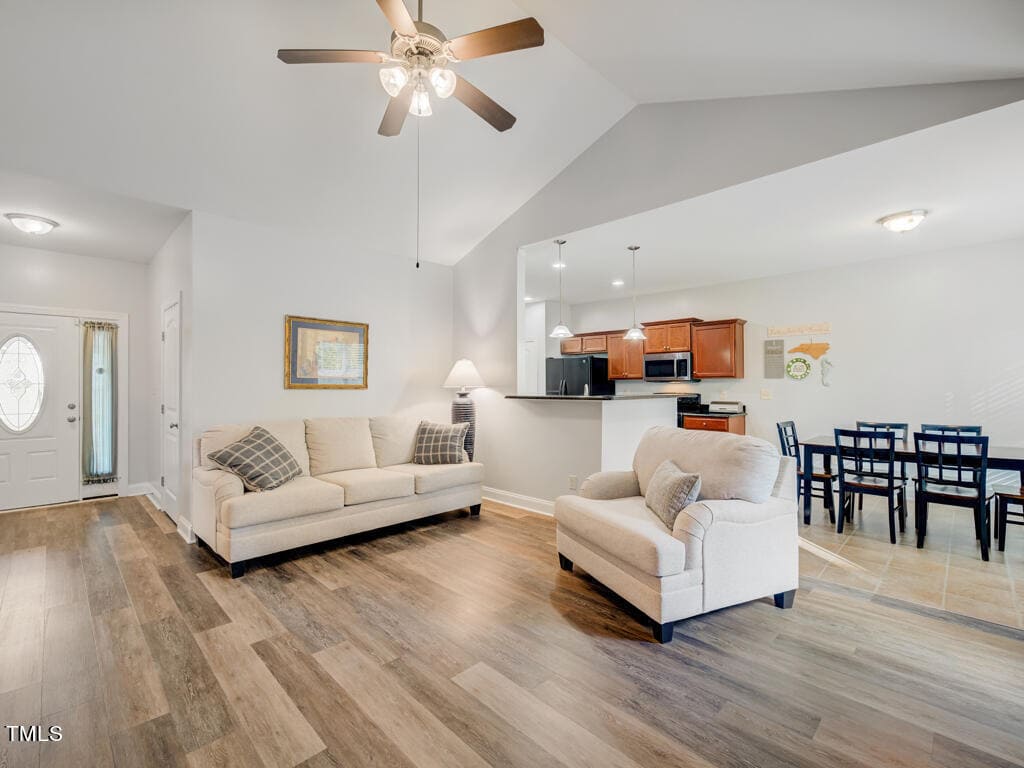 A spacious living and dining area with light wood flooring. There's a beige sofa and armchair, a ceiling fan, and a small dining table with chairs. The kitchen features wood cabinets and modern appliances. Walls are painted white with subtle decor.