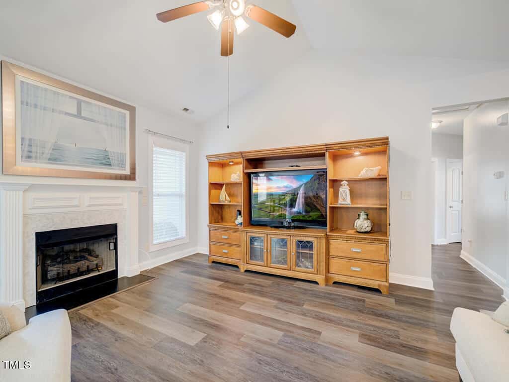 A cozy living room features a wooden entertainment center with a large flat-screen TV. A ceiling fan is overhead, and a fireplace with a framed ocean photo is on the left. The room has light-colored walls and wood flooring.