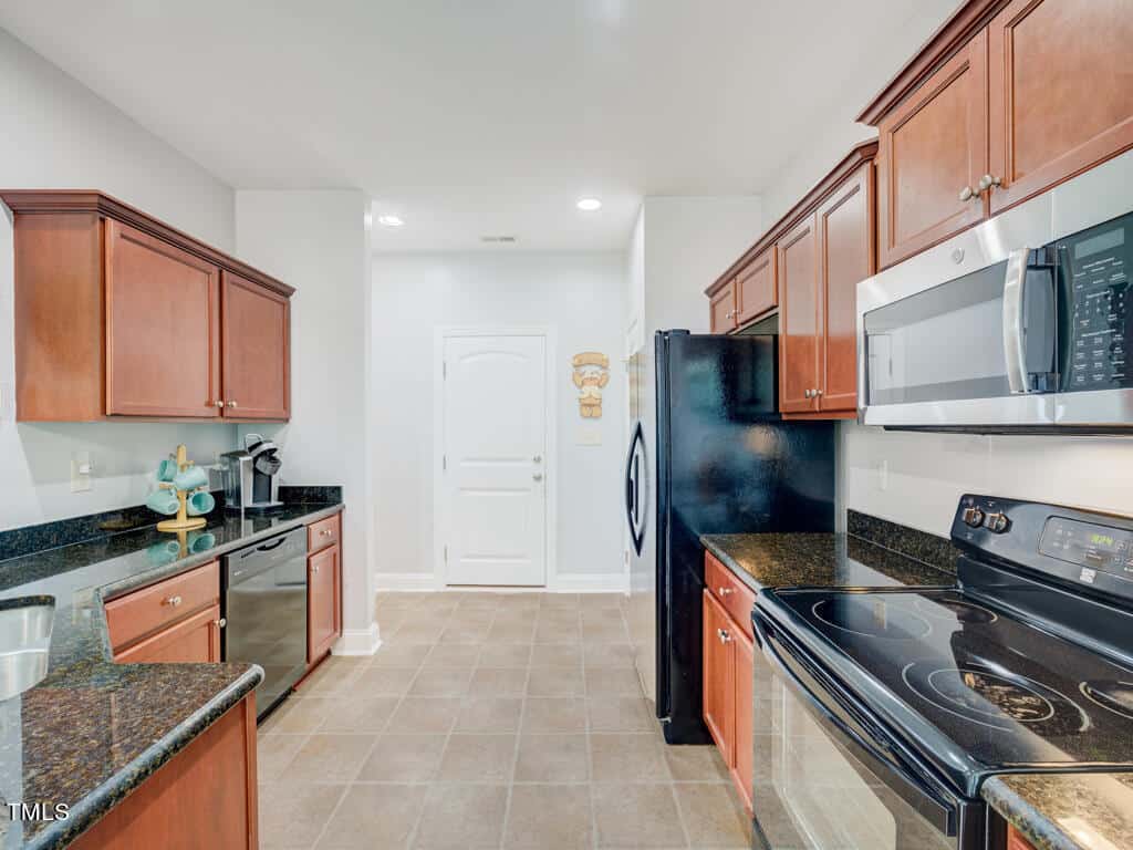 A modern kitchen with dark granite countertops, wooden cabinets, black appliances, and a stainless steel microwave. A coffee maker is on the counter. The floor is tiled, and a closed white door is at the back of the room.