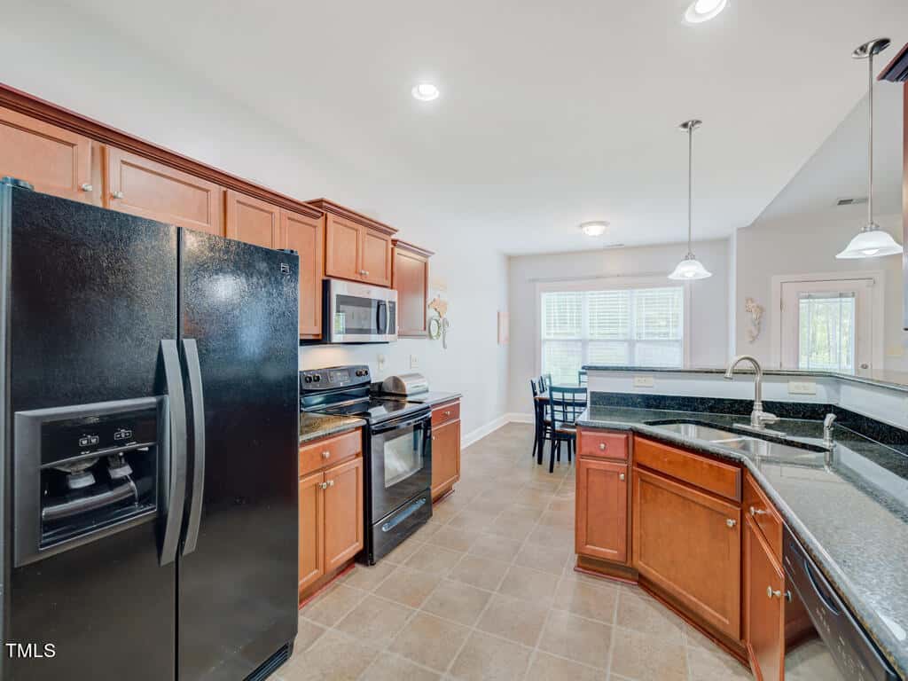 A spacious kitchen with wooden cabinets, black appliances, and a tiled floor. The room features a double-door fridge, oven, microwave, and a sink on a central island. A dining area with a table and chairs is visible in the background. Natural light streams in.