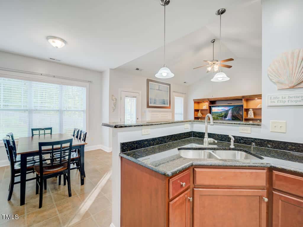 Bright, modern kitchen and dining area with dark granite countertops, wooden cabinets, and a double sink. A wooden dining table is set for six near large windows, and a ceiling fan and wall decor are visible in the background.