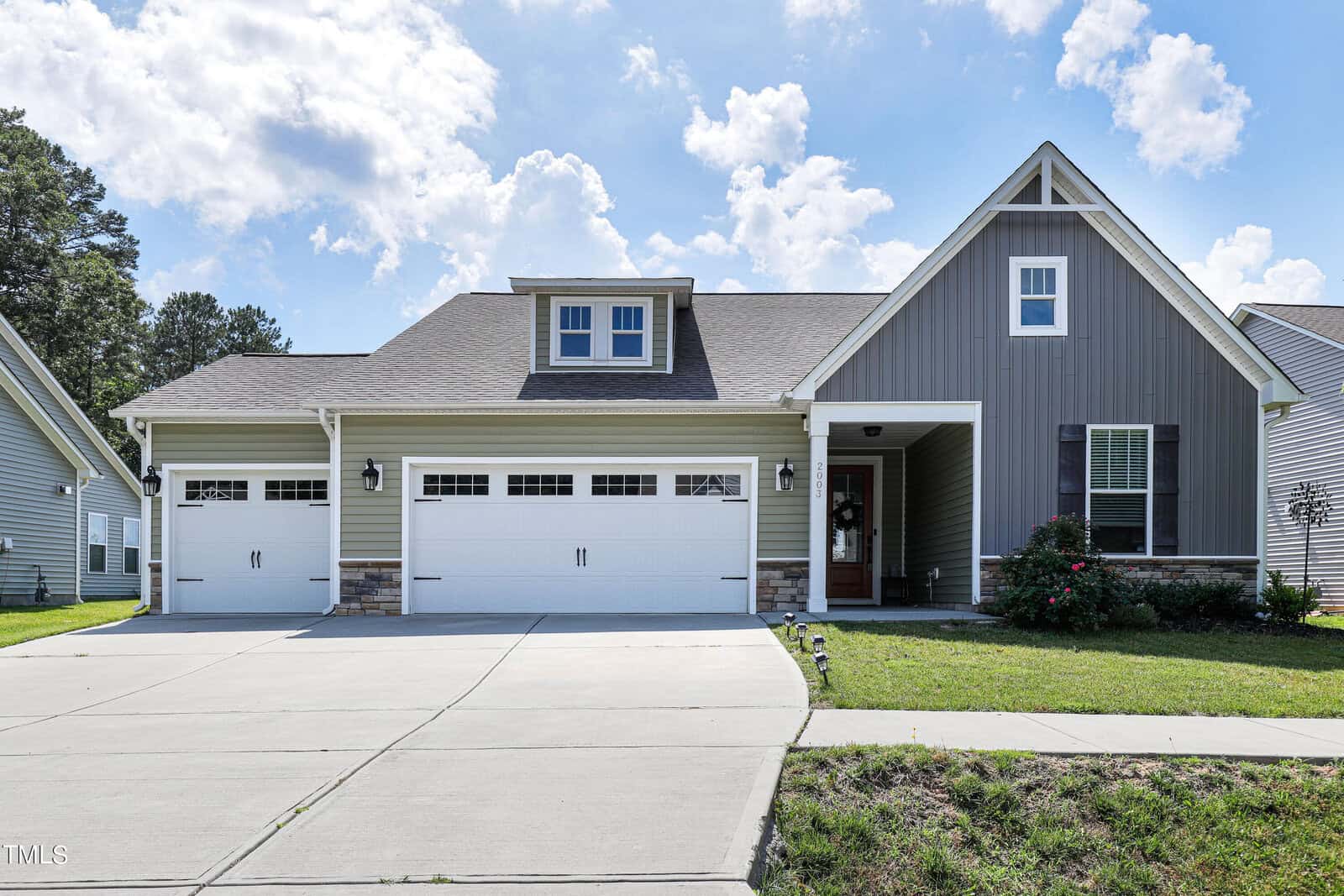 Located at 2003 Turning Leaf Lane in Creedmoor, this modern single-story house boasts a gray and white exterior. It features a three-car garage, a gabled roof, and a cozy porch. The spacious driveway is flanked by a lush lawn adorned with plants and shrubs under a partly cloudy sky.