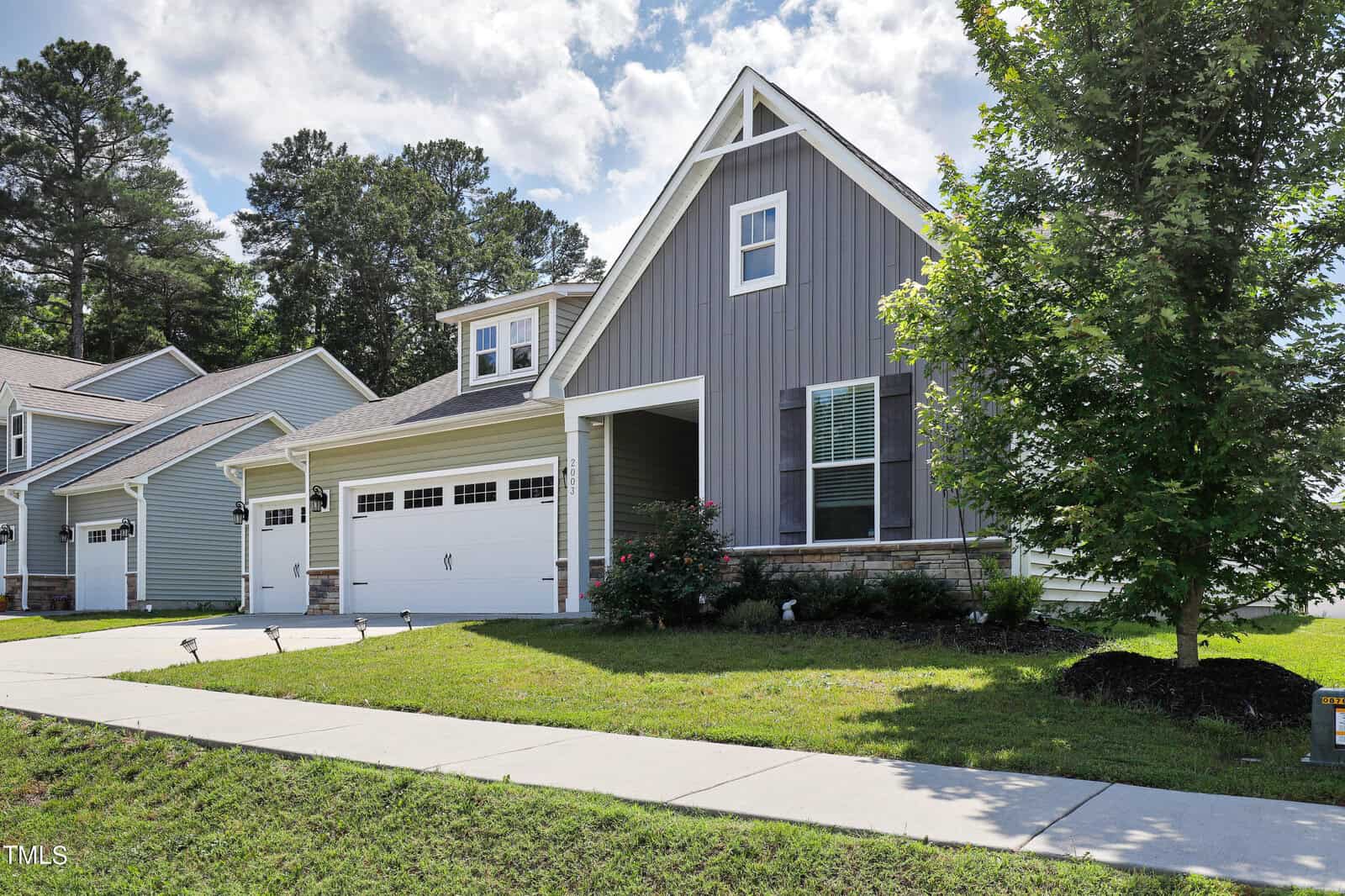 A modern suburban house with a gray facade, white trim, and a two-car garage. The front yard is landscaped with a small tree and bushes, and the sky is partly cloudy. Another house is visible on the left.